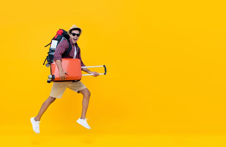 Smiling excited handsome Indian tourist man with backpack holding baggage and jumping ready to go for travel isolated on yellow background with copy space